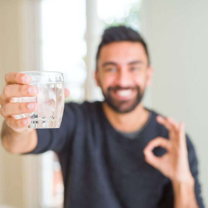 Hübscher hispanischer Mann, der ein frisches Glas Eauvation Wasser trinkt und mit den Fingern ein „OK“-Zeichen macht, ein ausgezeichnetes Symbol